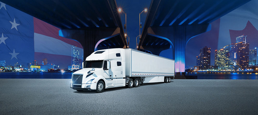 White tractor trailer parked under international bridge with Canada and USA flags and cityscapes in the background